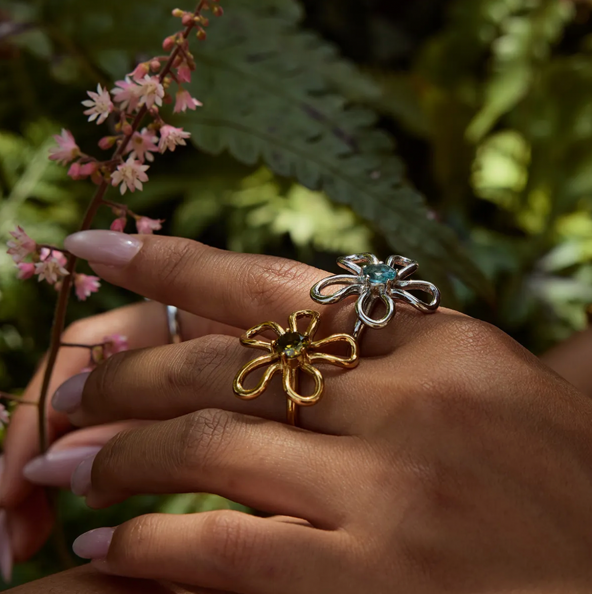 Close-up of a hand near pink flowers wearing the silver and gold wire-flower Vida RING styles side by side.