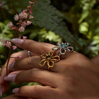 Close-up of a hand near pink flowers wearing the silver and gold wire-flower Vida RING styles side by side.