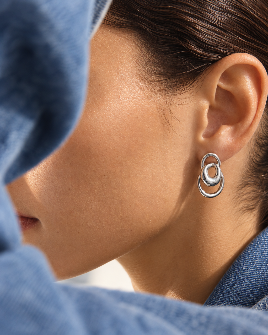 Silver interlocking-ring stud earring worn on a model's ear, styled with a denim hood — Folie à Trois Vortex EARRINGS.
