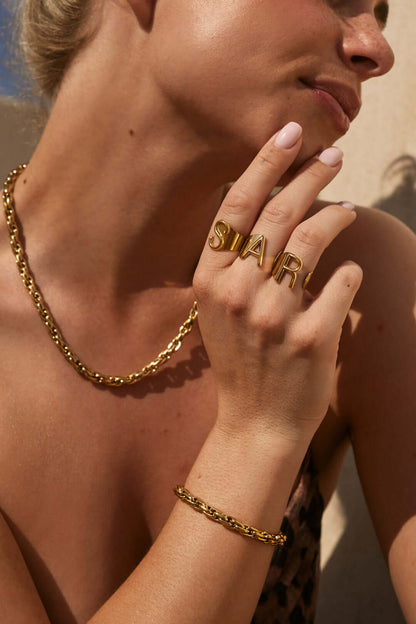 Close-up of a hand spelling out a name with stacked gold letter rings, worn near the chin in bright sunlight.