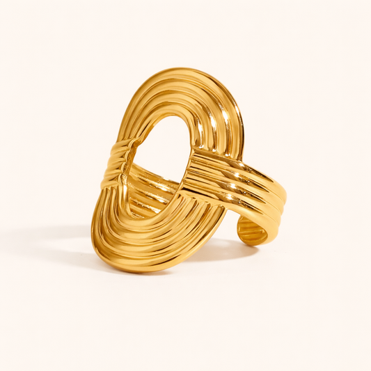 Ribbed gold ring with an open oval loop and textured wrapped band, shown at an angle on a cream backdrop — Folie à Trois Whisper RING.