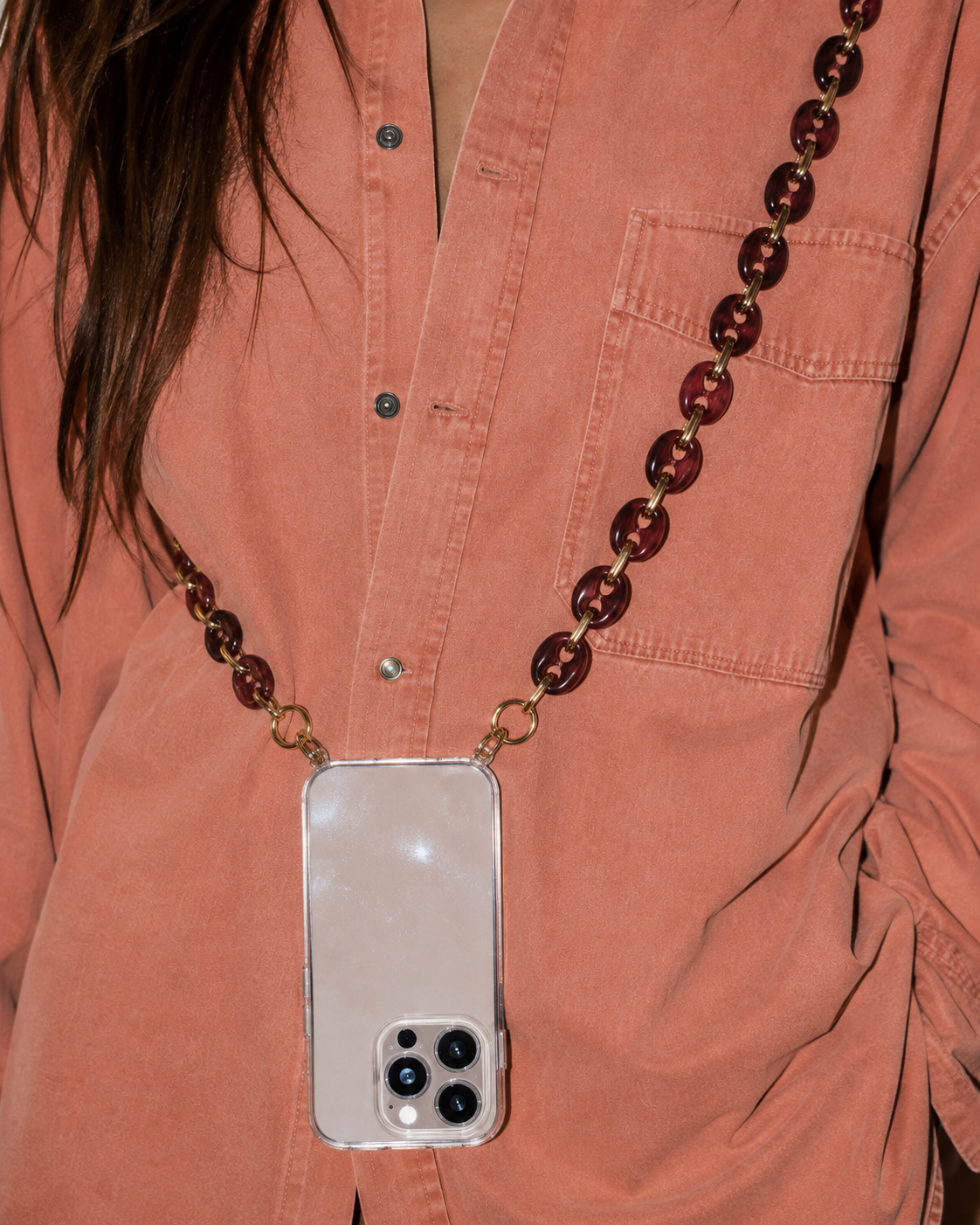 Model wearing a rust-colored shirt with a phone in a clear case suspended from the Folie à Trois Wilder wholesale phone chain in wine red puffed links.