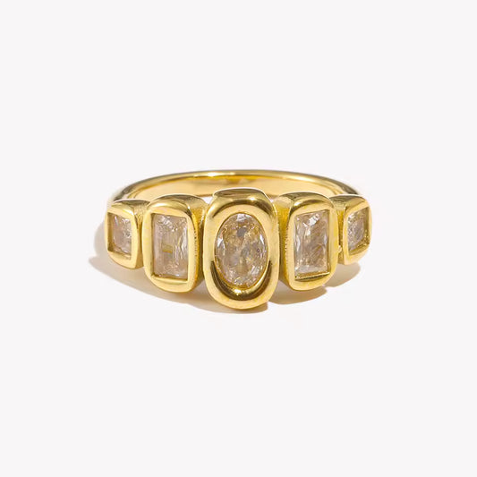 Gold ring set with five bezel-set clear stones of mixed oval and rectangular cuts in a row, shown on a cream backdrop — Zippora RING.