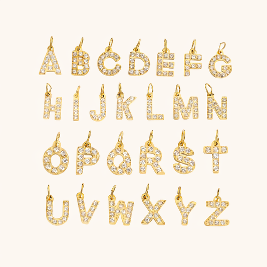The Zircon Letter CHARM collection in a flat lay grid, all 26 gold letters set with sparkling zircon stones on cream fabric.