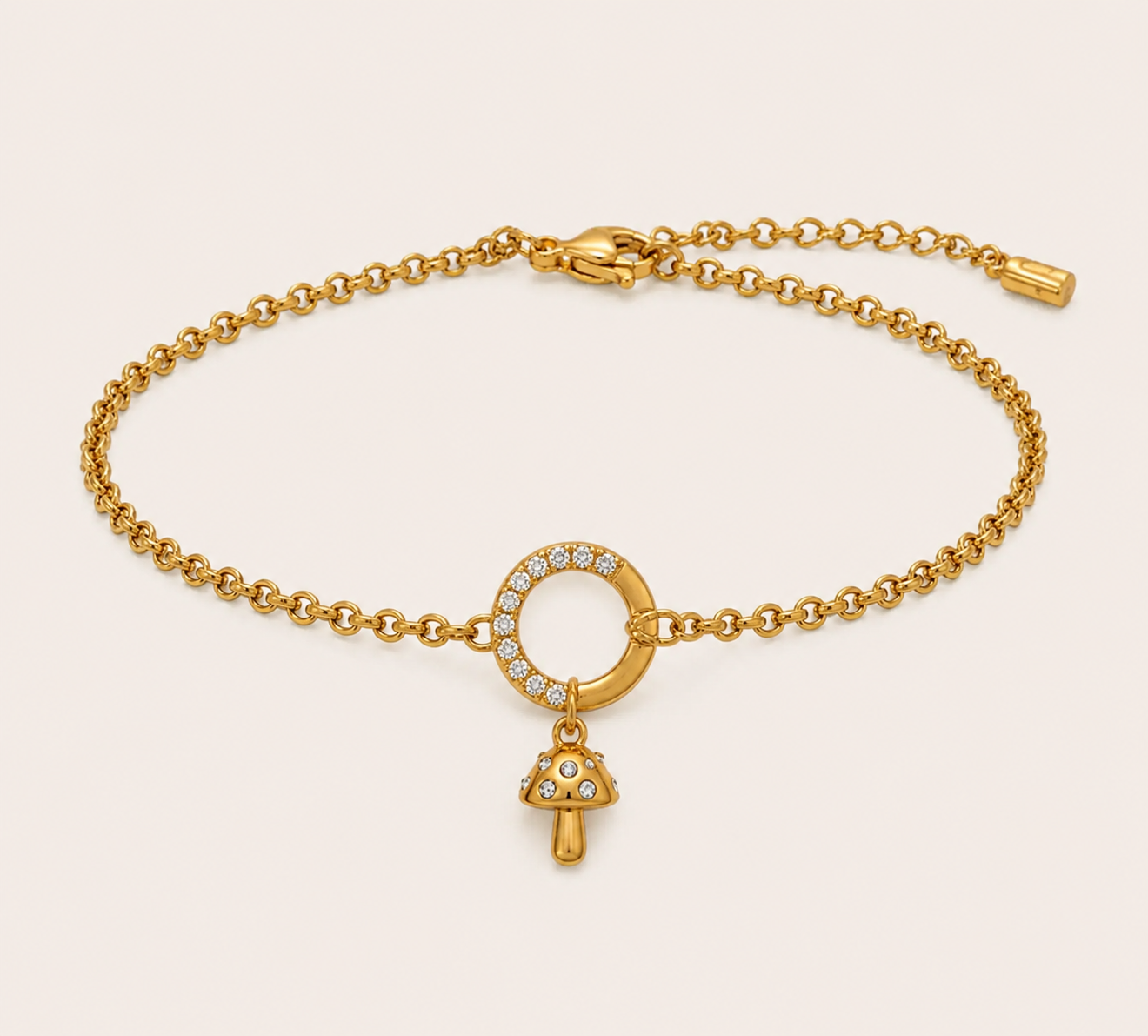 A gold chain bracelet with a circular ring set with small clear stones and a dangling mushroom-shaped charm, also accented with clear stones, shown on a light beige background.