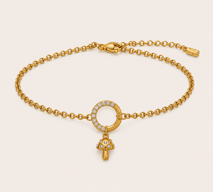 A gold chain bracelet with a circular ring set with small clear stones and a dangling mushroom-shaped charm, also accented with clear stones, shown on a light beige background.