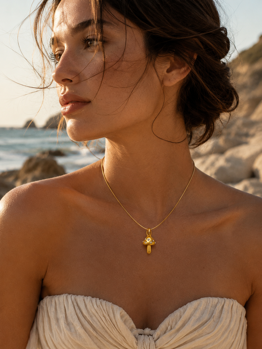 The Zircon Mushroom CHARM worn as a pendant on a fine gold chain necklace, resting at the neckline on a sunlit beach.