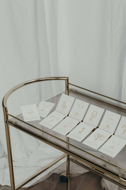 Twelve gold zodiac constellation stud earring cards laid out on a mirrored bar cart against a white curtain backdrop.
