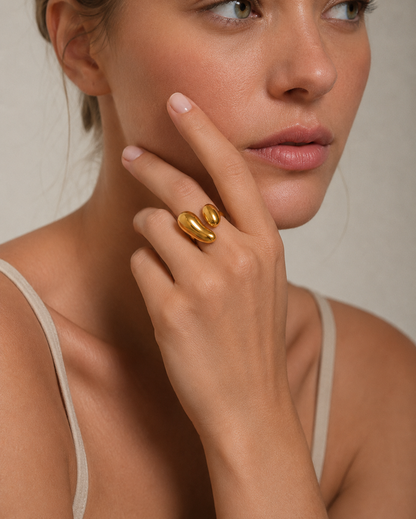 A close-up lifestyle shot of a model wearing a chunky, sculptural gold ring on her finger, with her hand resting against her cheek.