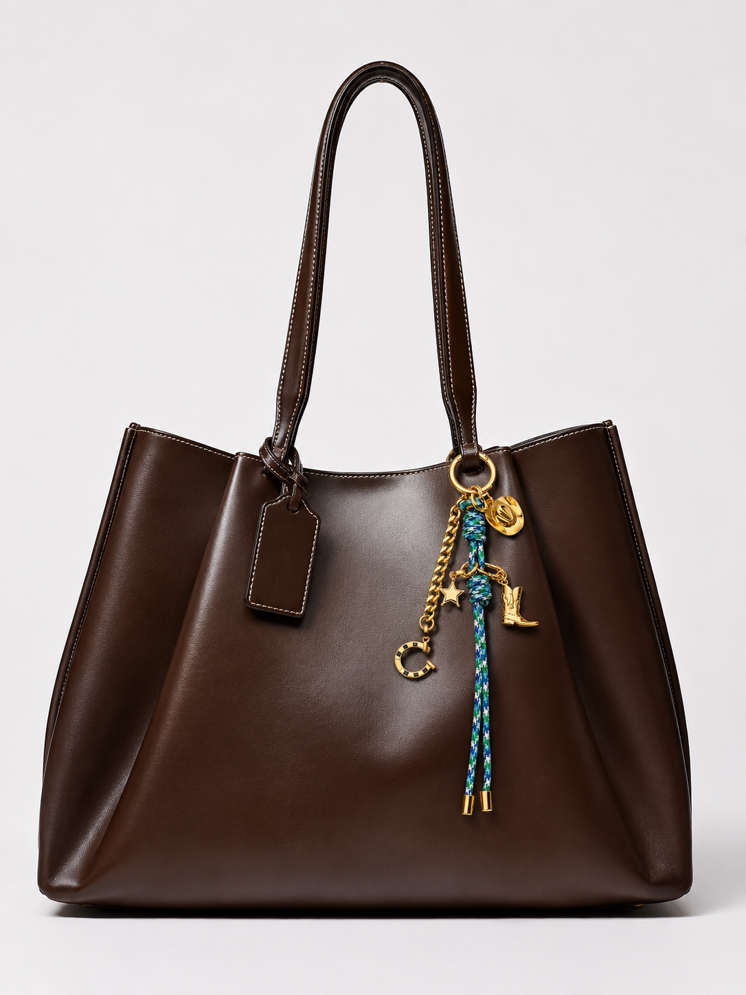 A dark brown leather tote bag with dual top handles, featuring a hanging gold-tone chain with a blue braided tassel and gold initials 'CL' charms, set against a plain light gray background.