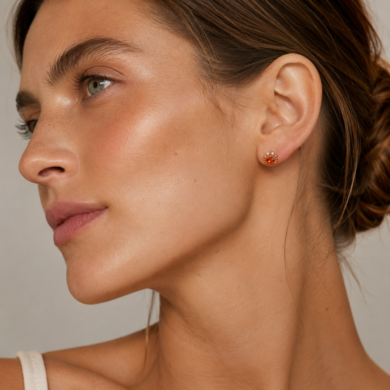 Close-up of a woman's ear wearing a small gold stud earring with a red gemstone, shown against a soft neutral background.