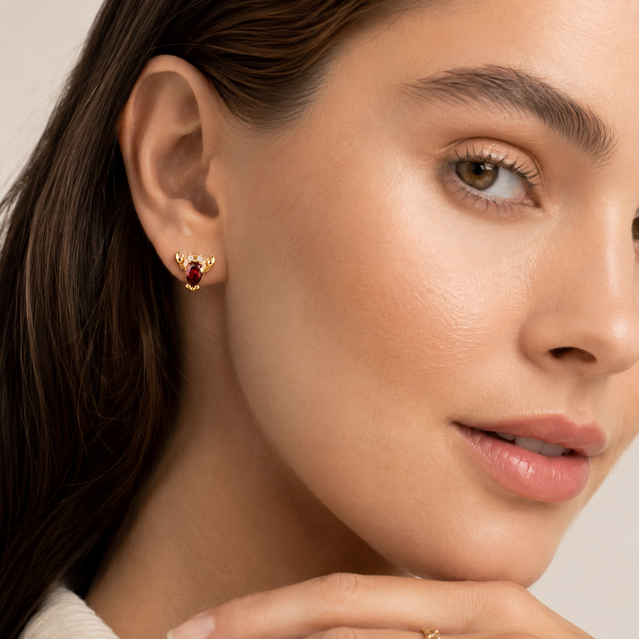 A close-up of a woman's ear wearing a small gold stud earring with a marquise-cut red gemstone, set against a soft beige background.