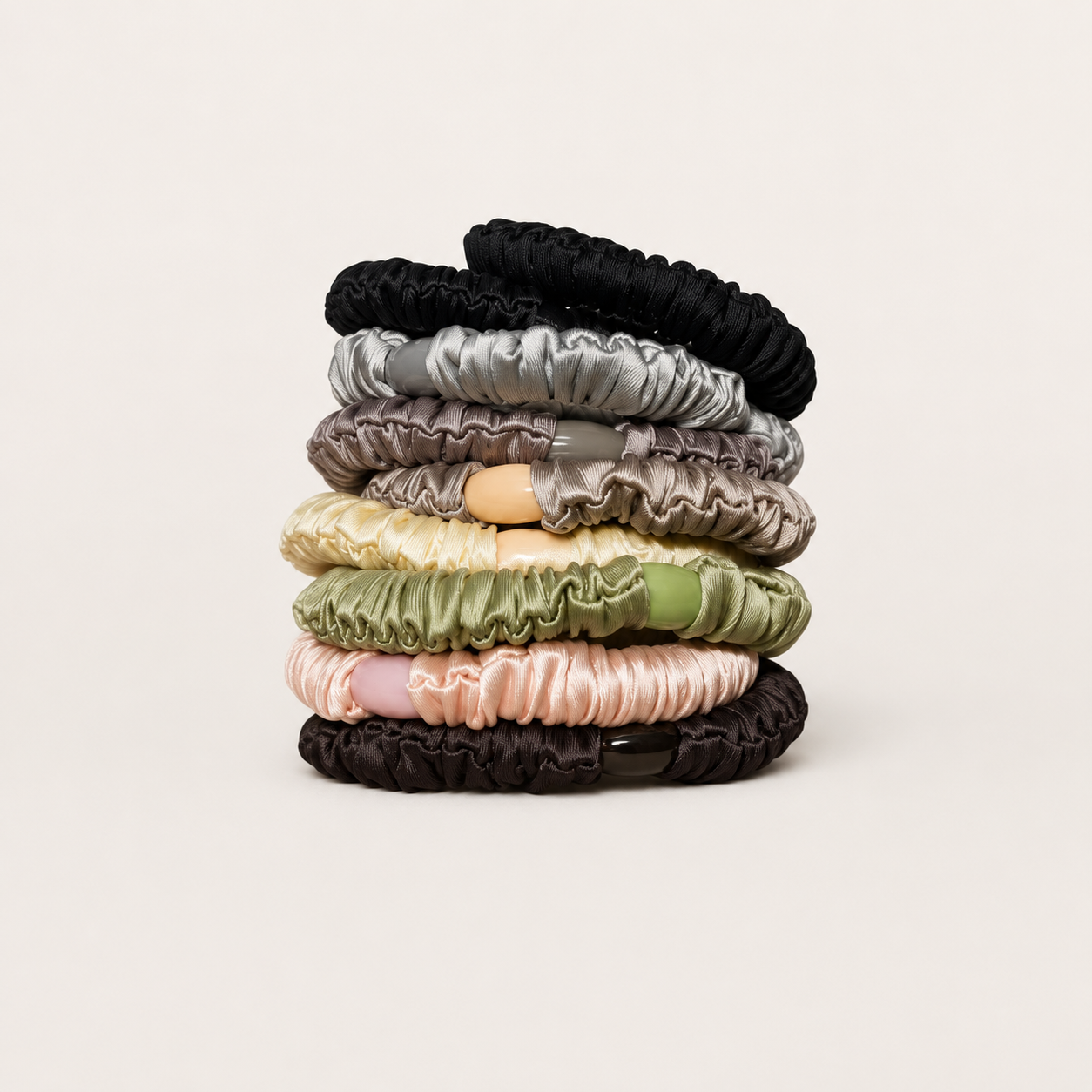 A stack of eight satin scrunchies in various colors including black, gray, taupe, cream, olive green, light pink, and dark brown, arranged on a light beige background.