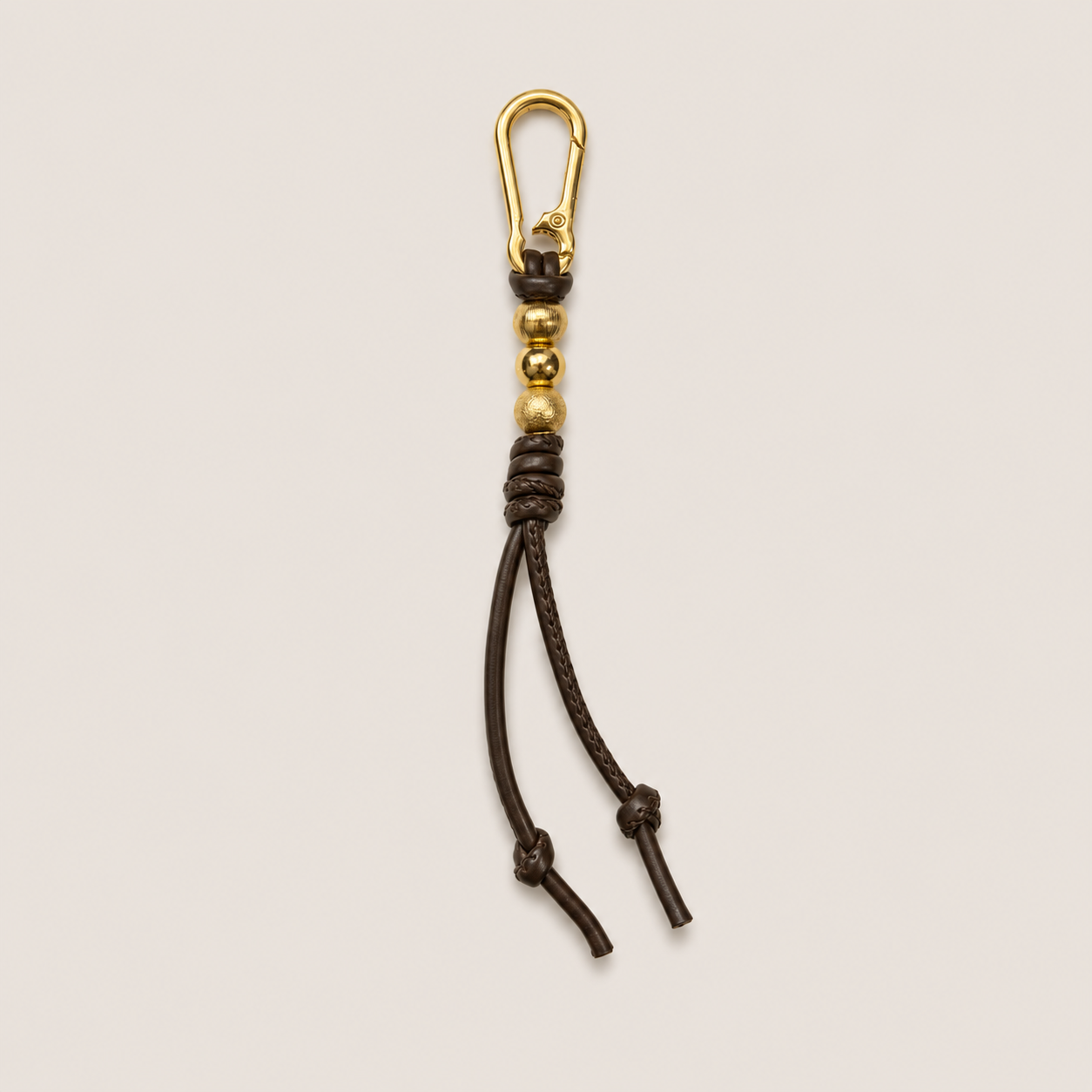 A dark brown leather keychain or bag charm with a gold-tone metal carabiner clip, decorative beads, and knotted tassels, shown on a light beige background.