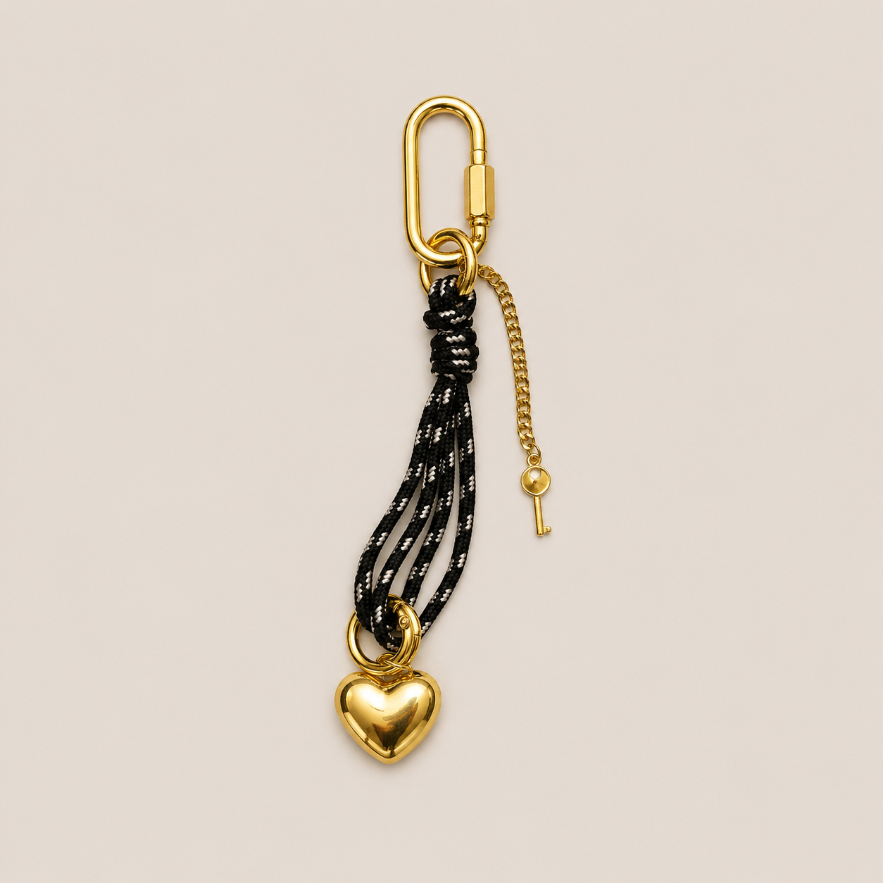 A gold-toned keychain featuring a carabiner clip, a short chain with a key, and a dangling heart-shaped charm attached to black and white braided cords, shown on a solid light beige background.