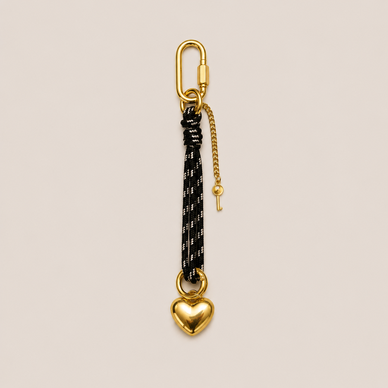 A gold carabiner keychain with a black and white patterned cord, a small gold chain with a key, and a gold heart-shaped clasp, shown on a light beige background.