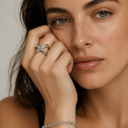 A close-up lifestyle shot of a woman wearing a silver infinity-style ring on her finger and a matching silver chain bracelet on her wrist, set against a soft, light beige background.