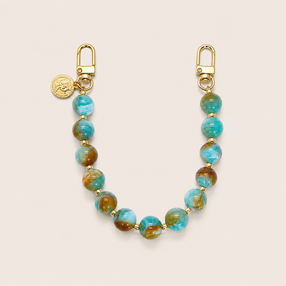 A beaded keychain or bag charm featuring round turquoise and brown marbled beads with gold-tone hardware, including lobster clasp ends and a small round gold coin charm, displayed on a light beige background.