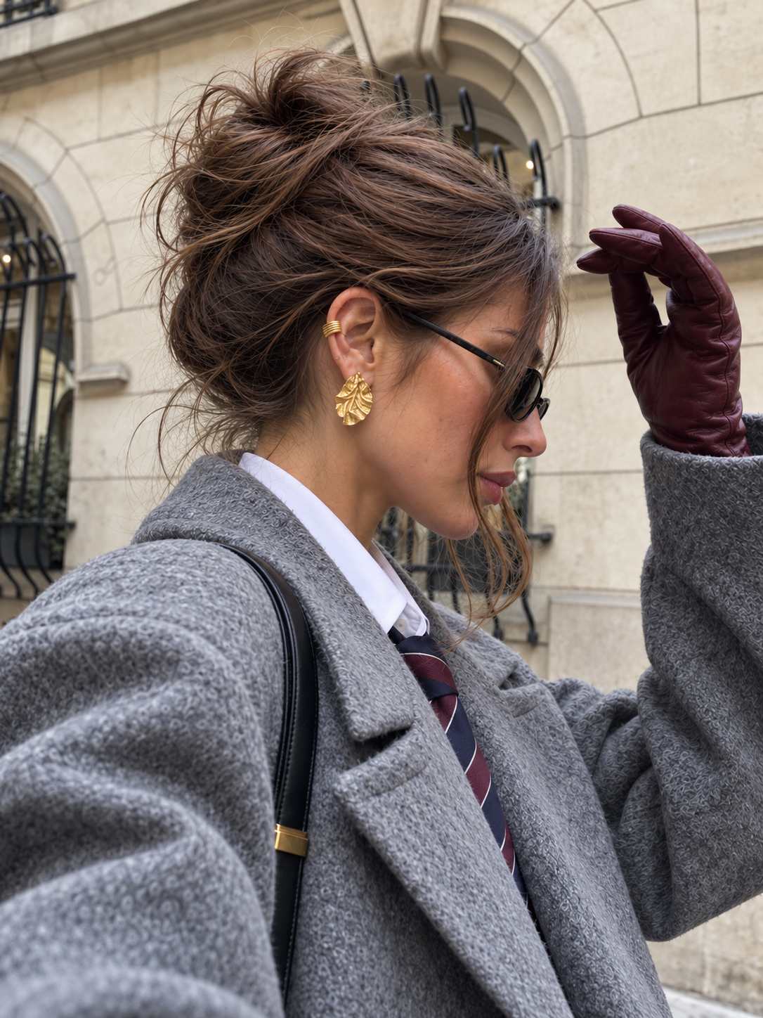 Woman with a messy bun, sunglasses and grey coat wears gold Feuille EARRINGS, ruffled leaf studs, on a city street — Folie à Trois