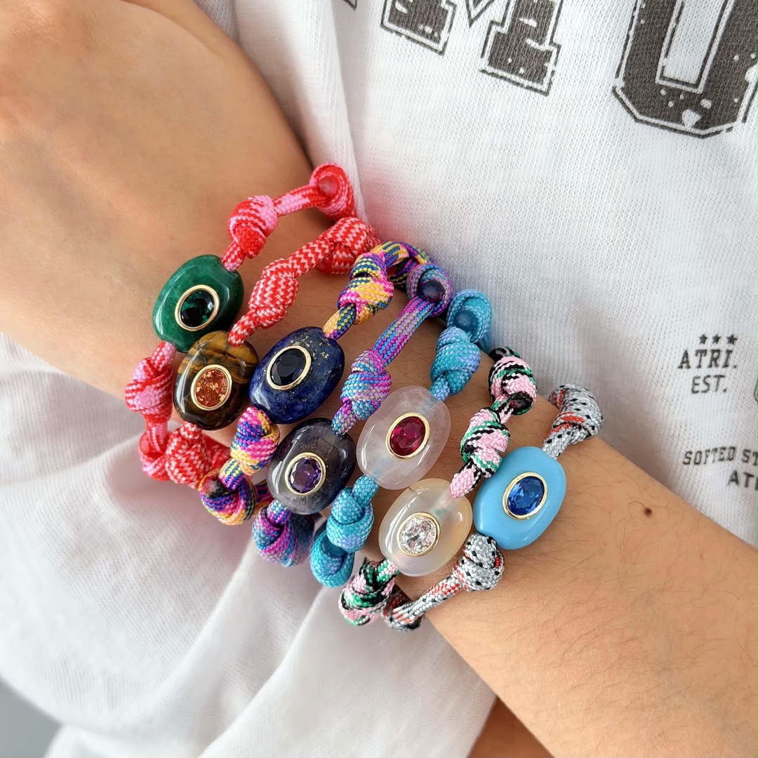 A close-up of a person's wrist wearing a stack of colorful beaded cord bracelets. The bracelets feature various colored beads, including green, blue, purple, pink, and white, with some displaying small gemstone-like centers.