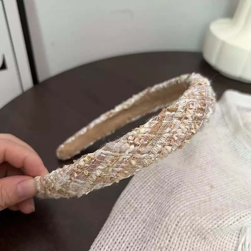 A single cream beige tweed woven headband held by hand on a dark wood table.