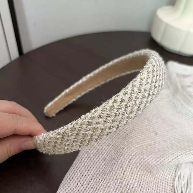 A single cream and gold metallic woven headband with sparkle thread held by hand on a dark table.