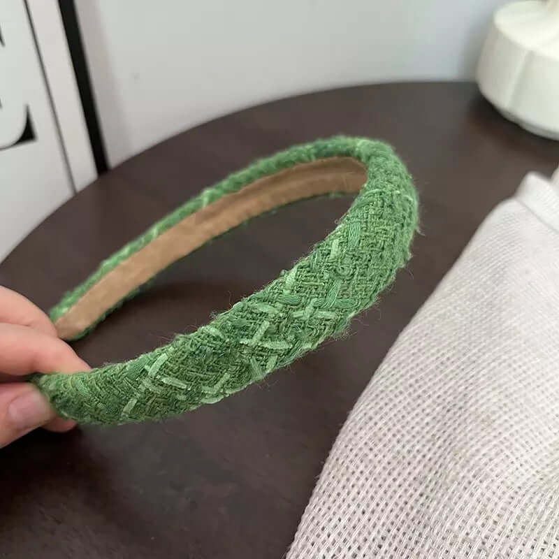 A single solid sage green woven tweed headband held by hand on a dark wood table.