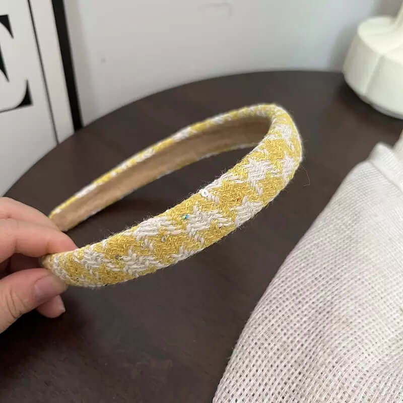 A single yellow and white houndstooth tweed headband held by hand on a dark wood table.