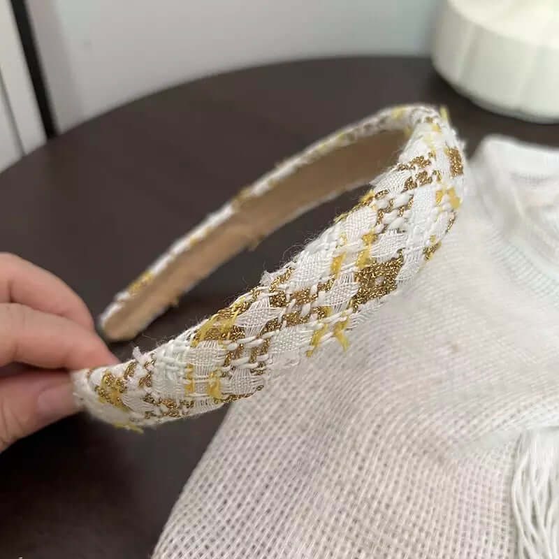 A single white and gold metallic diamond-weave tweed headband held by hand on a dark table.