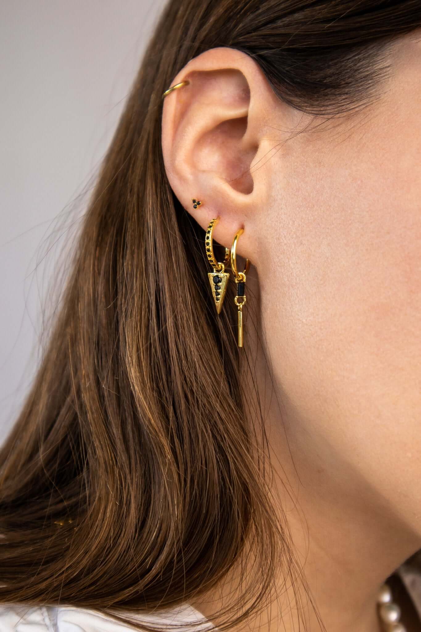 A model wears the Gabriella SET in gold with black onyx stones, layering the triangle dangle hoop and bar-dangle hoop along the ear.