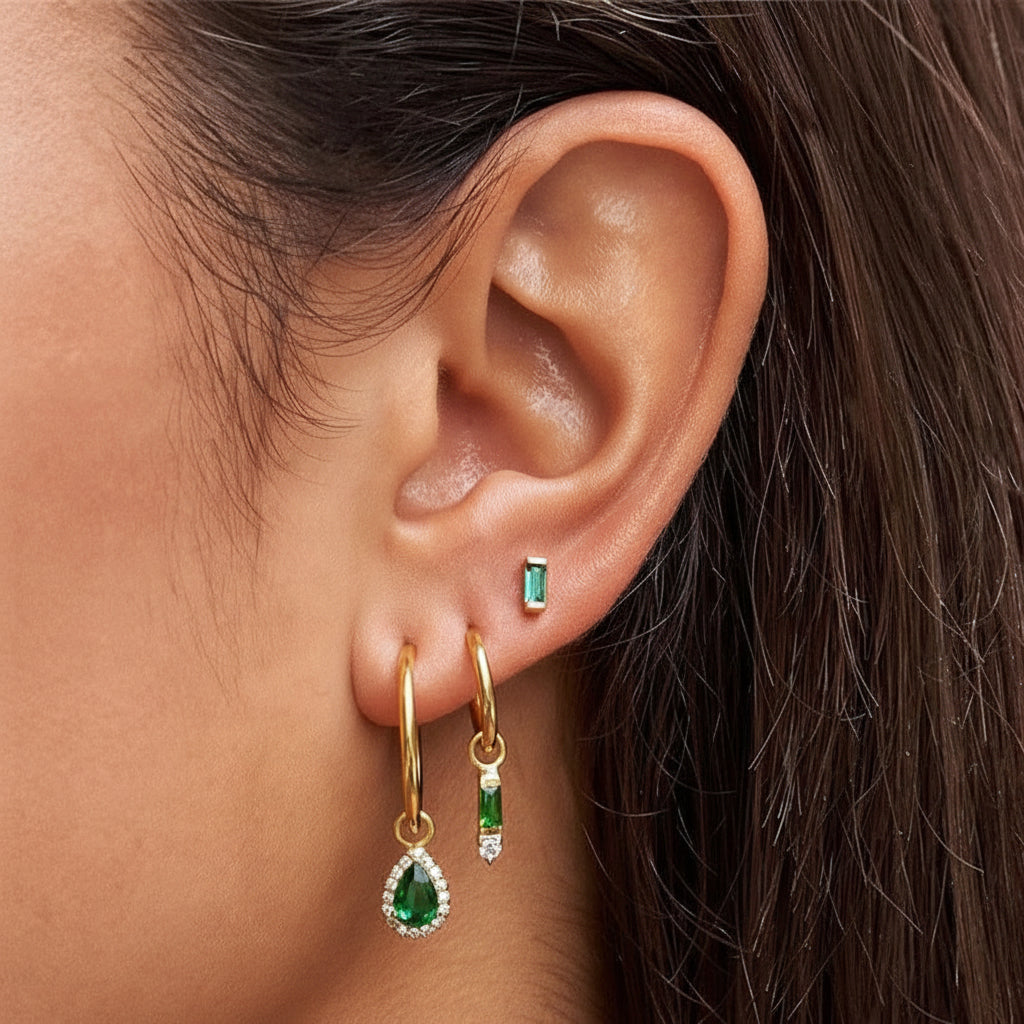 A model wears the Leila SET in gold with emerald green stones, layering the baguette stud with two dangle hoop earrings.
