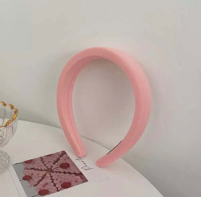 A single solid pink padded fabric headband with center stitching styled upright near a glass dish and photo card.