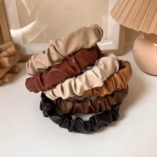 Group of five ruched faux leather Romy HAIRBAND headbands in beige, black, chestnut, cream and tan, styled near a picture frame and lamp.